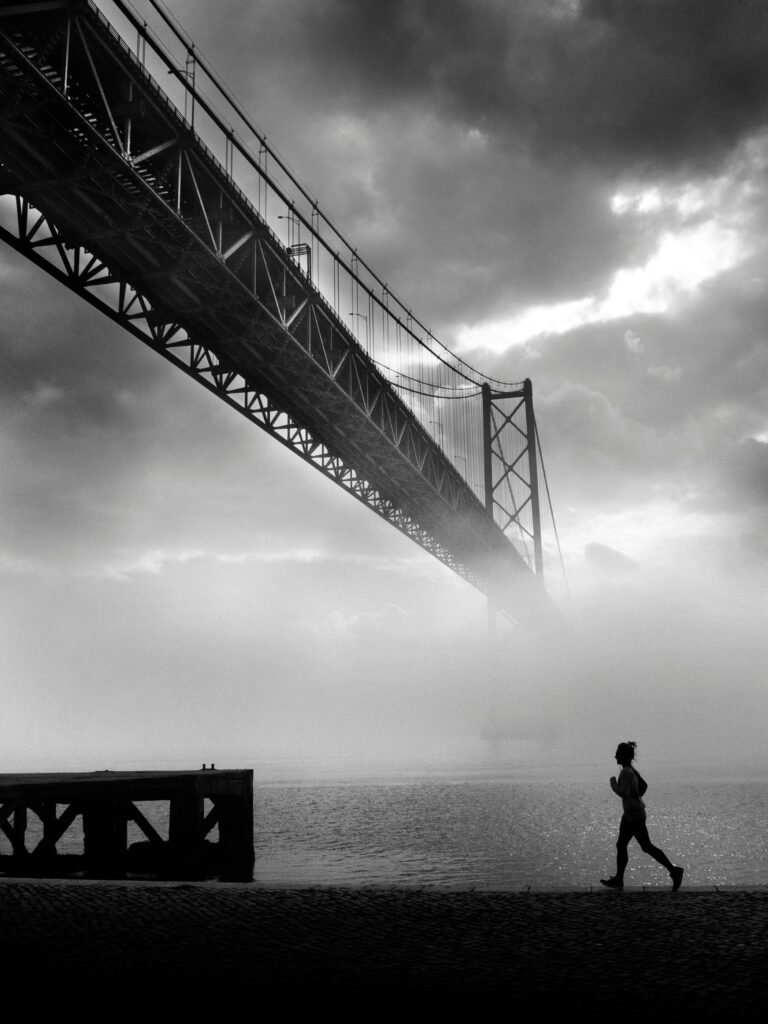 Silhouette of a person jogging near a misty river with a suspension bridge in the background.